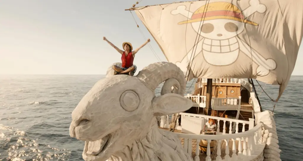 Luffy sentado na figura de proa do navio Going Merry no live-action de One Piece, com a bandeira dos Chapéus de Palha ao fundo.