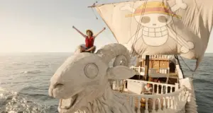 Luffy sentado na figura de proa do navio Going Merry no live-action de One Piece, com a bandeira dos Chapéus de Palha ao fundo.